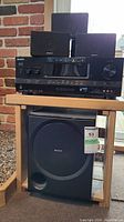 Front view of Sony STR-DH720 AV receiver with four satellite speakers arranged on and around it, and large Sony subwoofer below on a wooden stand against brick background.