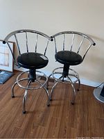 Pair of metal framed swivel bar stools with black vinyl round seats and chrome finish, placed on a wood floor against a beige wall.