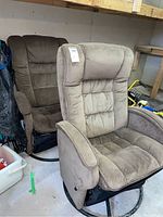 Photo showing two matching swivel glider chairs with brown faux suede upholstery, sitting side by side, in a garage setting. One chair is light brown and the other darker brown.