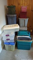 Photo showing stack of plastic storage containers and totes of various sizes and colors against wood-paneled wall.