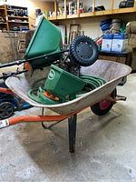 Metal wheelbarrow with red wheel and orange handles showing rust and heavy use, holding green hand-held spreader, Scotts push spreader, garden hose coil