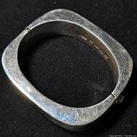 View of the square bracelet with rounded edges resting on a black background, showing the outer surface and the hinged clasp.