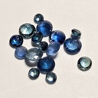 Collection of loose faceted blue sapphire gemstones in various cuts and shades of blue scattered on a white background.