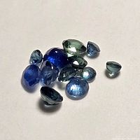Photo of 13 loose faceted blue sapphires of varying shades and cuts on a white surface showing color and clarity.