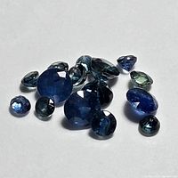 Multiple loose faceted blue sapphire gemstones in varying shapes and sizes on a white background, showing color and cut detail.