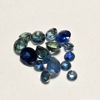 Loose blue sapphire gemstones of varying sizes and shapes on white background, showing mixed colors of blue and green tones.