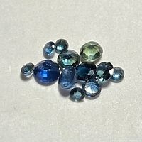 Top view of twelve loose blue sapphires in mixed cuts and sizes laid on a light surface showing color variation and good clarity.