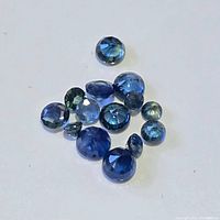 Loose blue sapphire gemstones of varying size and cuts lying on a white surface, showing deep blue to bluish-green colors and faceted shapes.