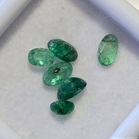 Close-up photo of six loose oval emerald gemstones in a white display box showing green hues and natural inclusions.
