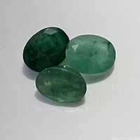 Three oval cut emerald gemstones in varying shades of green on white background, front view.