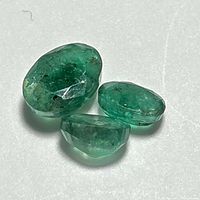 Three loose emerald gemstones placed on white surface showing front and side views to highlight clarity, cut, and color.