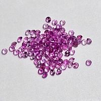 Close-up image of numerous small, round-cut natural pink sapphires arranged on a white background, showing their pink coloration and faceted surfaces.