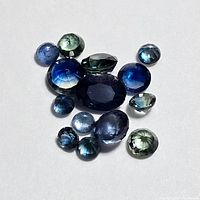Top-down close-up photo of 14 loose sapphires in varied shapes, sizes, and colors including blue and green hues on a white background.