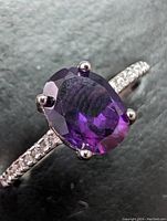 Close-up image of a sterling silver ring featuring a faceted oval natural amethyst gemstone with four prongs and a band set with small clear stones.