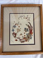 Full view of the framed dried pressed flower artwork showing the whole piece with a wood frame and double matting in cream and blue with colorful pressed flowers arranged in a wreath design.