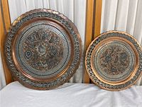 Two copper tone etched metal plates displayed side by side on a white surface with a curtain background.