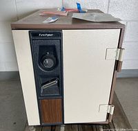 Front and side view of a sturdy metal combination safe with beige door and brown panels, showing the lock dial and handle section with woodgrain accent.