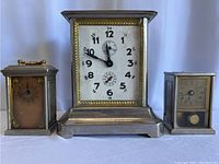 Front view of three clocks: larger metal clock in center with white face and two smaller brass clocks on either side.