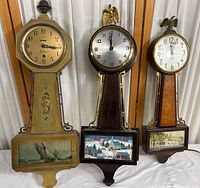Three banjo clocks arranged side by side, showing differences in size, design, and decoration.