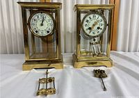 Front view of two brass table clocks with glass panels, measuring about 6.5x5.5x11 inches, with pendulum parts placed in front.
