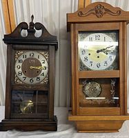 Two vintage wood wall clocks side by side on a white surface, one dark wood Veritas clock with gold-tone dial accents and one lighter Ansonia clock with floral carved top and etched glass panel.