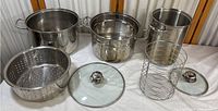Wide view of all items in the set showing large steamer pot, tall asparagus steamer basket, perforated steaming insert, wire basket, and glass lids.