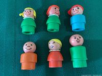 Six vintage Fisher Price Little People wooden figures displayed on green fabric surface, showing front view of faces and bodies in various colors.