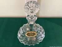 Overall view of the clear Baccarat crystal cognac bottle with stopper showing its fluted shape and ornate stopper on top, hanging on a green cloth background.