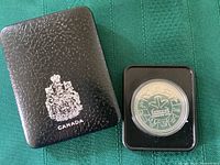 Black textured presentation case with silver-embossed Canadian coat of arms next to coin in plastic protective capsule showing the 1978 Edmonton Commonwealth Games design.