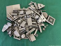 Photo shows a variety of dark grey Lego pieces scattered on a green fabric background including plates, bricks, and small elements.