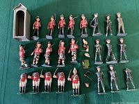 Photo showing assortment of approximately 25 lead soldiers and a small guard house structure. Figures mostly in red coats with black trousers and bearskin hats, varied poses and some weaponry visible. Loose parts and accessories also laid out.