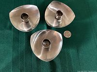 Three stainless steel candle holders arranged on green fabric with a quarter for scale. Each has a smooth, rounded triangular form with a cylindrical socket for taper candles in the center.