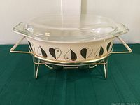 Full view of Pyrex casserole dish in golden hearts pattern on warming stand with clear lid.