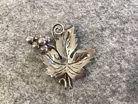 Sterling silver leaf brooch with a five-lobed grapevine leaf, attached grape cluster of polished silver balls, and a curled tendril. Displayed on grey fabric background.