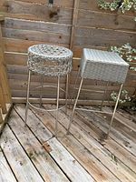 Two unique bar stools side by side on wooden deck floor in front of wood fence. One round woven seat with metal legs, one square woven seat with metal legs.