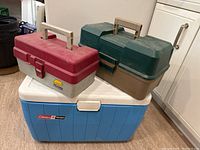 Photo showing two stacked fishing tackle boxes (red/gray and green/brown) on top of a blue Coleman cooler with white lid in a kitchen setting.