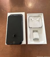 Apple iPhone 7 with cracked screen placed on top of original box showing accessories EarPods and charged USB adapter inside box.