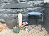 Photo of six garden planters of varying shapes, sizes and materials alongside a small blue metal table on a wooden deck against a gray stone wall.