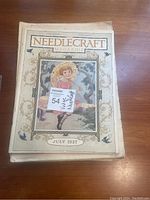 Front cover of July 1927 Needlecraft Magazine showing vintage illustrated artwork of a young girl in a pink dress sitting on a garden wall with flowers.