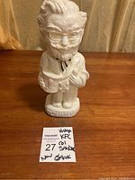 Front view of white ceramic Colonel Sanders bank on wood surface with handwritten note 'Vintage KFC Col Sanders Bank'.