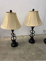 Two beige fabric shade table lamps with black twisted metal bases on a concrete floor against a white wall.