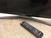 Close-up image of the Samsung TV logo at the bottom center of the TV and the Samsung remote control placed on the floor in front of the base.