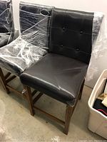Four black upholstered dining chairs with button-tufted backs and wooden legs, some wrapped in protective plastic, viewed from side angle.