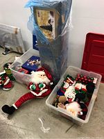 Artificial Christmas tree boxed and wrapped in plastic, placed on floor next to plastic bins and a large plush Santa decoration