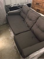 Photo showing the entire grey fabric couch wrapped partially in protective plastic, positioned next to a wooden dresser and a black box.