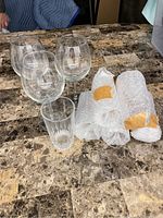 Photo of four stemmed wine glasses, one tall ribbed tumbler glass, and four additional glasses wrapped in bubble wrap on a marble surface.