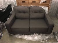 Full front view of dark gray fabric love seat sofa showing two seat and two back cushions with tufted buttons, wide armrests, wrapped partially in plastic for protection.