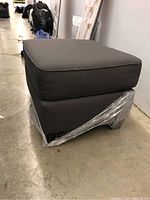 Side view of dark gray fabric ottoman with plastic wrap on base.