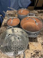 Set of three copper interior pots of varying sizes with glass lids and silver metal handles, along with a perforated stainless steel steamer insert with handles, placed on a granite countertop.