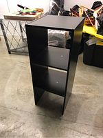 Full view of a black, narrow bookshelf with three shelves on a concrete floor surrounded by other household items.